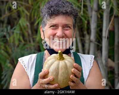 Anziani felice positivo caucasico fattoria donna giardiniere con capelli ricci tiene una zucca organica con entrambe le mani e sorrisi per lo spettatore. Foto Stock