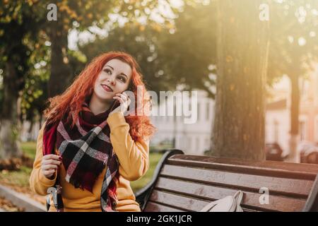 Giovane donna dai capelli rossi in felpa gialla che parla al telefono. Lady siede sul banco del parco il giorno dell'autunno. Stile di vita. Foto Stock