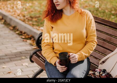 Ritratto di giovane donna dai capelli rossi seduta in parco su panca con tazza di caffè. La donna indossa una felpa gialla e tiene una tazza riutilizzabile. Stile di vita Foto Stock