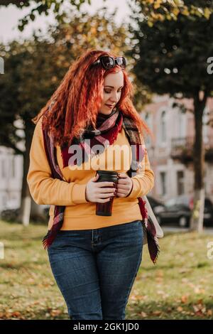 Giovane donna dai capelli rossi in maglione giallo e occhiali da sole tiene la tazza termica con il caffè nelle sue mani. Lady passeggiate attraverso la città e il parco su autunno soleggiato da Foto Stock