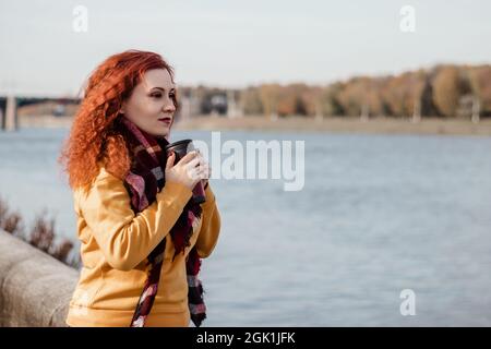 Giovane donna dai capelli rossi beve caffè sul lungomare e gode di vista sul fiume. La donna indossa una felpa gialla e tiene una tazza termica riutilizzabile. Eco-fr Foto Stock