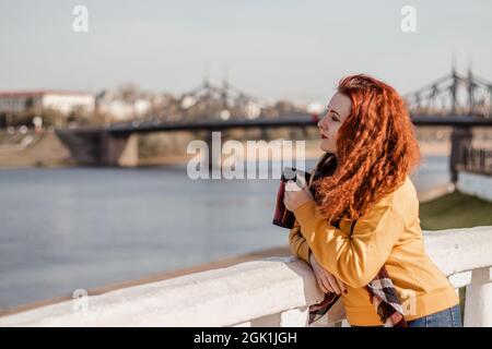 Giovane donna dai capelli rossi in maglione giallo tiene in mano la tazza termica con il caffè. Goditi la vista sul fiume nella soleggiata giornata autunnale. Stile di vita ecologico, Foto Stock