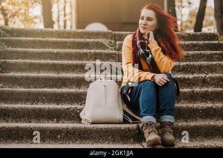 Giovane donna dai capelli rossi siede sulle scale nel parco. Lady in maglione giallo sta godendo giorno di sole. Turismo urbano. Stile di vita, spazio di copia. Foto Stock