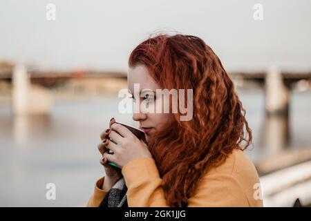 Ritratto di giovane donna dai capelli rossi sullo sfondo del fiume e del ponte della città. Beve caffè da una tazza di carta e gode di una vista autunnale. Spazio di copia. Foto Stock