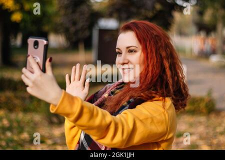 Giovane donna dai capelli rossi sta videochattando. Utilizza uno smartphone moderno. La signora sorride e cammina nel parco in una giornata d'autunno soleggiato. Foto Stock