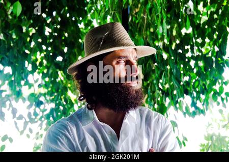 il giovane uomo con barba e capelli lunghi indossa cappello e camicia bianca circondata dal verde Foto Stock