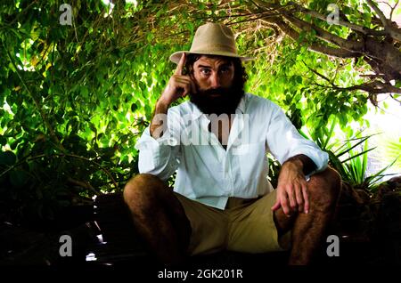 il giovane uomo con barba e capelli lunghi indossa cappello e camicia bianca circondata dal verde Foto Stock