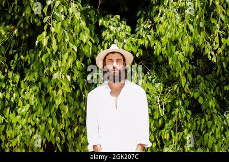 il giovane uomo con barba e capelli lunghi indossa cappello e camicia bianca circondata dal verde Foto Stock