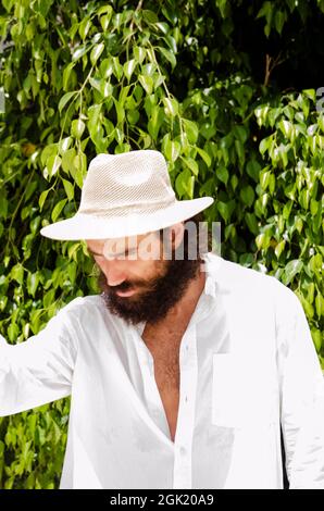 il giovane uomo con barba e capelli lunghi indossa cappello e camicia bianca circondata dal verde Foto Stock