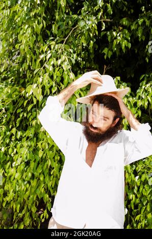 il giovane uomo con barba e capelli lunghi indossa cappello e camicia bianca circondata dal verde Foto Stock