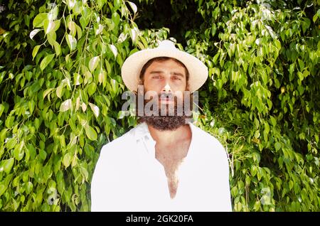 il giovane uomo con barba e capelli lunghi indossa cappello e camicia bianca circondata dal verde Foto Stock