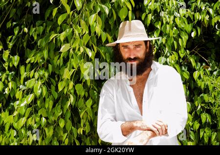 il giovane uomo con barba e capelli lunghi indossa cappello e camicia bianca circondata dal verde Foto Stock