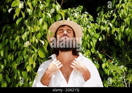 il giovane uomo con barba e capelli lunghi indossa cappello e camicia bianca circondata dal verde Foto Stock