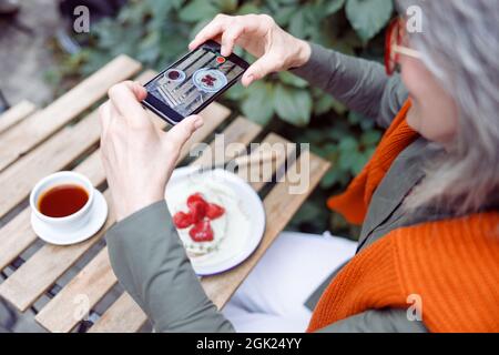 Ospite anziano scatta foto del dessert alle fragole con smartphone al tavolo all'aperto Foto Stock