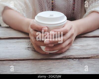 Chiudi le mani femminili tenendo una tazza di carta da caffè bianca su un tavolo di legno. Giovane donna che beve caffè da tazza usa e getta. Foto Stock