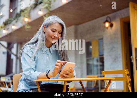 Donna asiatica superiore positiva surfs internet con telefono seduto al tavolo giallo Foto Stock