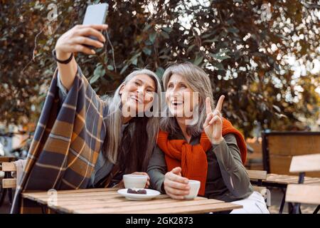 Le donne anziane gioiose prendono selfie con smartphone seduti a un piccolo tavolo in Street cafe Foto Stock