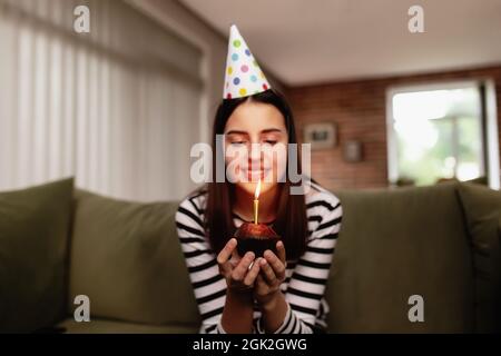 La brunetta della ragazza di compleanno in cappello di cartone siede sul divano o sul divano e soffia fuori la candela che brucia sul cupcake. Donna che tiene la torta di compleanno nelle sue mani. C Foto Stock