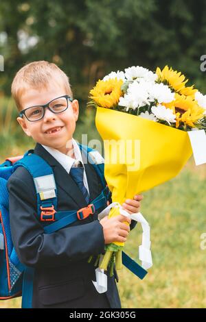 Piccolo sorridente ragazzo con fiori pronto ad andare in prima classe a scuola. Uno scolaro in uniforme scolastica e in uno zaino si erge sulla strada con un bou Foto Stock