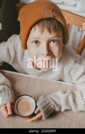 un ragazzo in un maglione e un cappello marrone beve il cacao da una tazza rossa. Una foto accogliente con una tazza in mano. Ragazzo con occhi grandi guarda dal basso, vista dall'alto Foto Stock