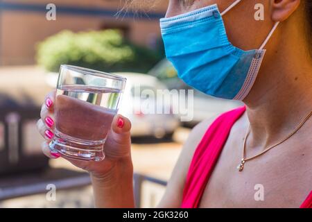 Ragazza in blu maschera medica all'aperto tiene in mano un bicchiere di acqua ancora pura. Bere alcolici nel periodo post pandemico. Indossare una maschera protettiva Foto Stock