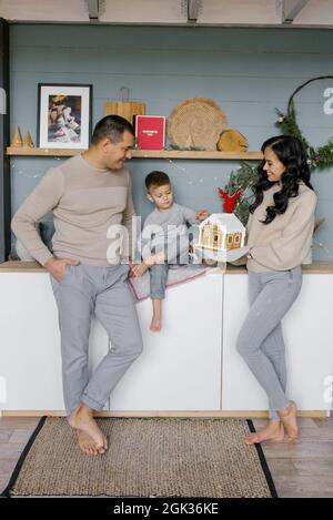 Una giovane ed elegante famiglia con un bambino piccolo sta in piedi nella loro cucina, decorata per Natale e ammirando una casa di pan di zenzero preparata con loro Foto Stock