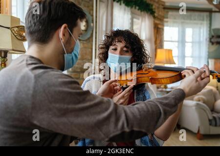 La donna felice suona il violino sotto le istruzioni dell'insegnante di musica in maschera durante il coronavirus a casa Foto Stock