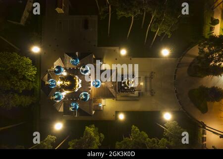 Vista aerea della chiesa illuminata tra i pini di notte. Nuova cappella pubblica vuota bianca con cupole dorate e blu all'aperto dall'alto. Bel luogo religioso, sacro per i ministeri Foto Stock
