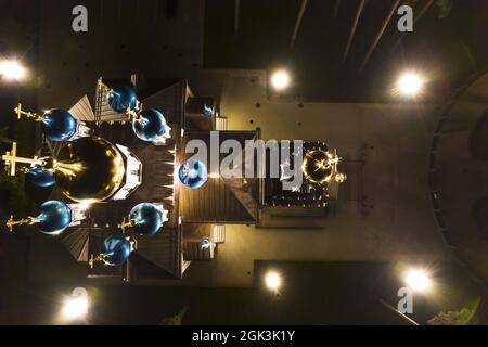 Vista aerea della chiesa illuminata tra i pini di notte. Nuova cappella pubblica vuota bianca con cupole dorate e blu all'aperto dall'alto. Bel luogo religioso, sacro per i ministeri Foto Stock