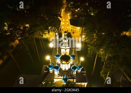 Vista aerea della chiesa illuminata tra i pini di notte. Nuova cappella pubblica vuota bianca con cupole dorate e blu all'aperto dall'alto. Bel luogo religioso, sacro per i ministeri Foto Stock