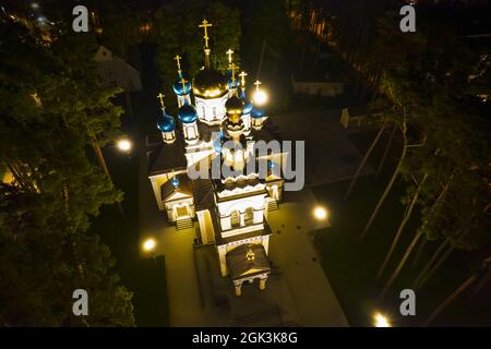 Vista aerea della chiesa illuminata tra i pini di notte. Nuova cappella pubblica vuota bianca con cupole dorate e blu all'aperto dall'alto. Bel luogo religioso, sacro per i ministeri Foto Stock