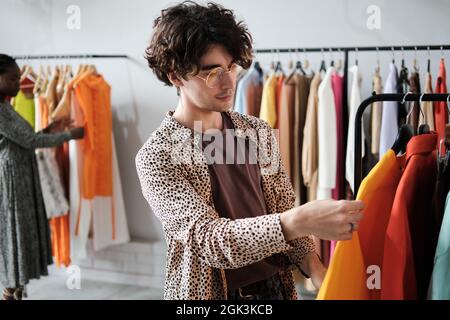 Giovane uomo alla moda che sceglie nuovi vestiti sul rack e che fa acquisti nel centro commerciale Foto Stock