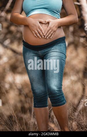 Pancia di una donna incinta che fa il cuore forma con le sue mani su di esso. Attività sane all'aperto. Trascorrere una calda giornata d'autunno nel parco. Nuovo concetto di vita. Foto Stock