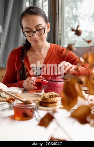 La donna che indossa un maglione arancione versa un tè caldo da una teiera rossa in una tazza di vetro sul tavolo. Miele, biscotti e libri vicino alla tazza di tè sul lino t Foto Stock