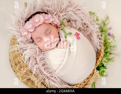 Ritratto di bella neonato ragazza swaadled in tessuto e vestendo la corona con fiori che dormono in cestino con pelliccia durante studio photoshoot. Carino Foto Stock