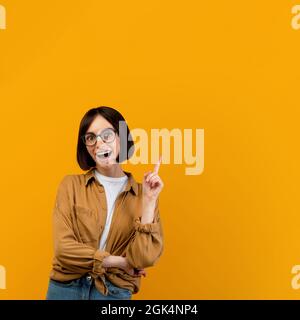 Giovane donna eccitata che punta il dito verso l'alto sullo spazio di copia e guarda la fotocamera isolata su sfondo giallo Foto Stock