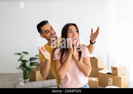 Eccitato ragazzo arabo sorprende la sua ragazza caucasica, che si sposta a casa propria insieme, in piedi in casa tra le scatole Foto Stock