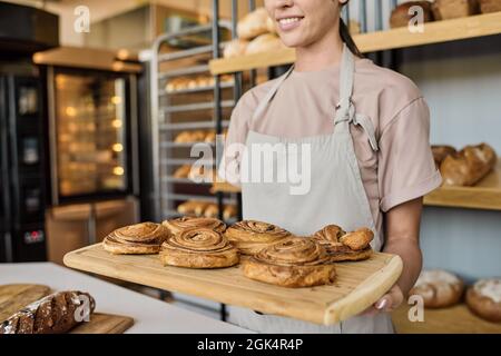 Mani di giovane panettiere femmina che tiene vassoio di legno con pasticceria fresca appetitosa a esposizione con pane Foto Stock