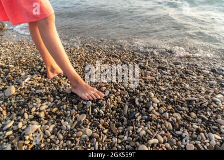 Donna a piedi nudi si erge su una spiaggia di ciottoli vicino a calme acque marine Foto Stock