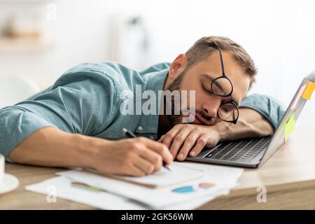 Uomo da millenario esausto che dorme sulla scrivania dell'ufficio, accanto a computer portatili e documenti, stanco di lavorare troppo Foto Stock