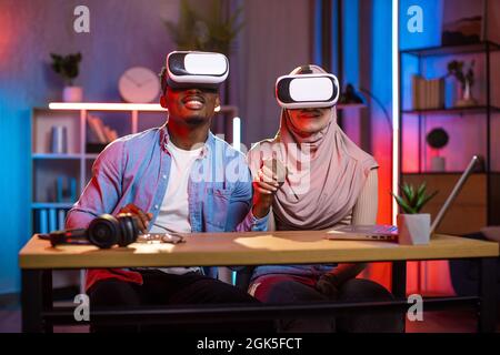 Donna araba e uomo africano seduti alla scrivania e tenendo le mani indossando cuffie per realtà virtuale. I giovani si divertono con la nuova tecnologia alla moda a casa. Concetto di gioco. Foto Stock
