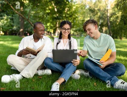 Allegri studenti multirazziali che parlano e usano un computer portatile, che riposano nel campus universitario, spazio libero Foto Stock