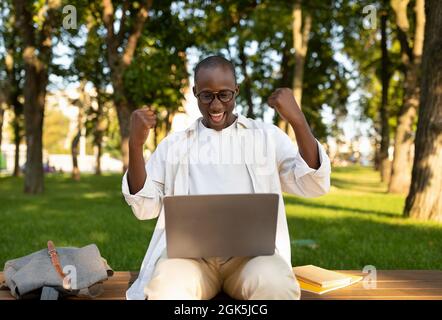 Concetto di istruzione remota. Ragazzo nero molto gioioso con computer portatile gesturing sì all'aperto, seduto nel campus universitario Foto Stock