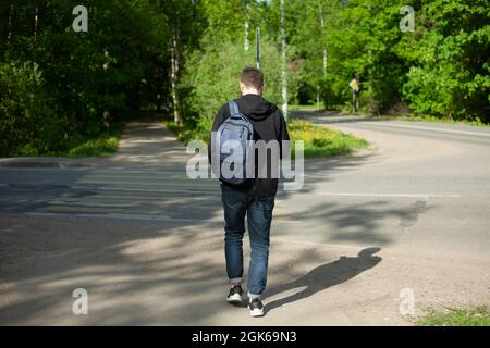 Un ragazzo con uno zaino cammina lungo la strada. L'uomo sulla strada pedonale. L'uomo dalla parte posteriore. L'uomo va al lavoro. Foto Stock