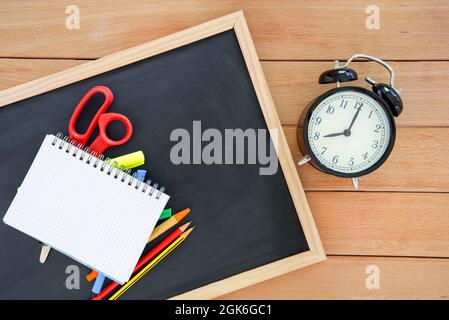 Notebook aperto e colorati suthies scuola su una piccola lavagna adagiata su un tavolo di legno vicino ad una sveglia. Spazio di copia. Concetto di ritorno a scuola. Foto Stock