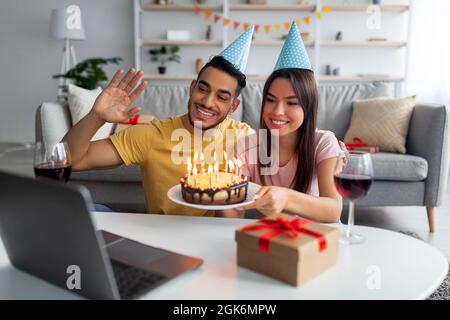 Festa di compleanno online. Coppia multirazziale con videochiamata, mostrando torta con candele al laptop webcam da casa Foto Stock