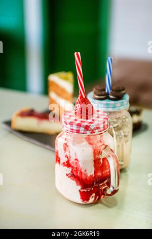 Frullati di fragole e dietro cioccolatini in bicchieri di vetro con coperchi in metallo, su un tavolo verde e con diversi pezzi di torta sullo sfondo. Foto Stock