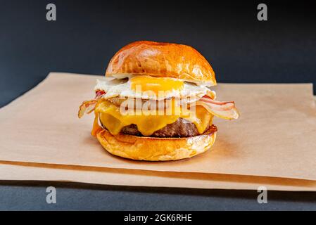 Grande hamburger di manzo su un lucido con uova fritte, pancetta affettata, formaggio Cheddar sciolto gocciolante, cipolla bianca cotta a fette su carta marrone greaseproof Foto Stock