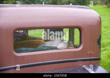 Una donna anziana in cappello e impermeabile sta guidando in vecchia auto retrò d'epoca in colori marroni. Grandmather con ombrello dietro la finestra rettangolare posteriore della vecchia automobile antica sotto la pioggia Foto Stock