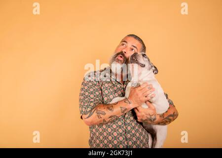 Un ritratto di studio di un uomo barbuto che tiene il suo cane sul suo braccio con un volto stupito Foto Stock